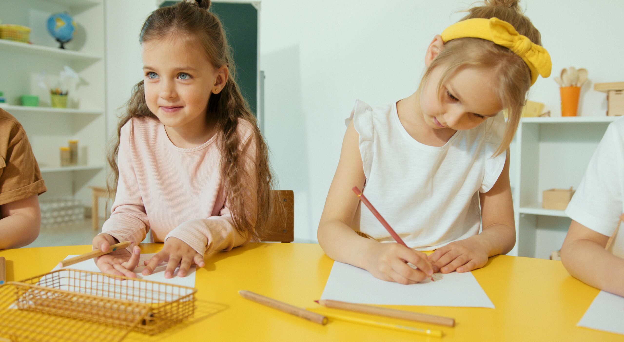 Two young girls writing and learning in a bright classroom setting.